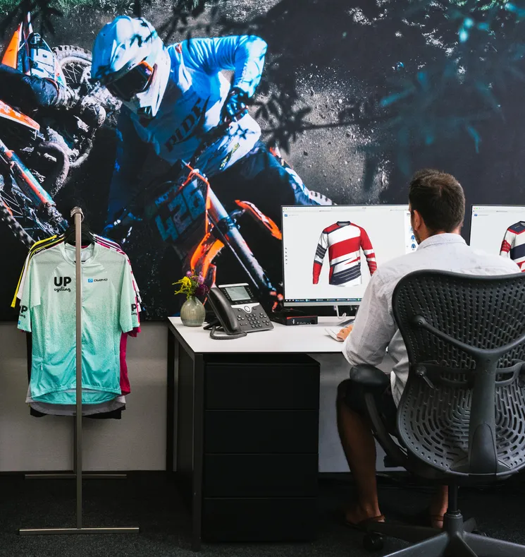 A man sits at a desk designing a customized sports jersey on a computer, with a large image of a motorcyclist and a clothing rack with various sportswear in the background.