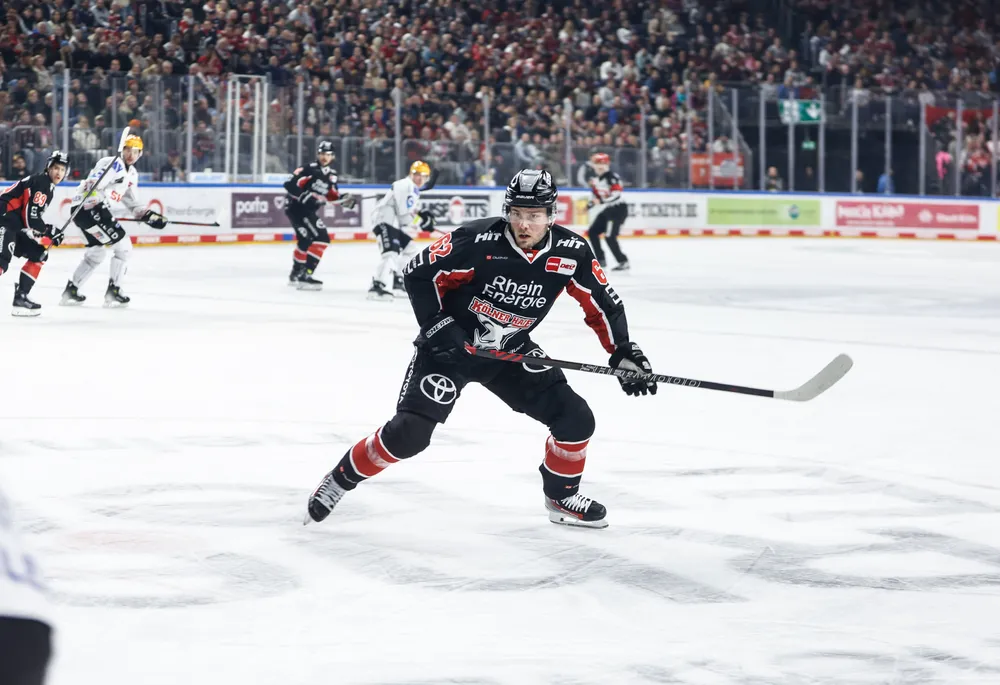 An ice hockey player wearing a black jersey with red and white accents skating on the ice with a stick.
