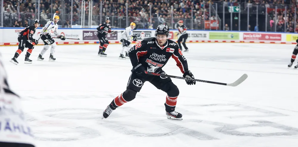 An ice hockey player in a black jersey with red and white accents skating on the ice with a stick, while spectators cheer in the background.
