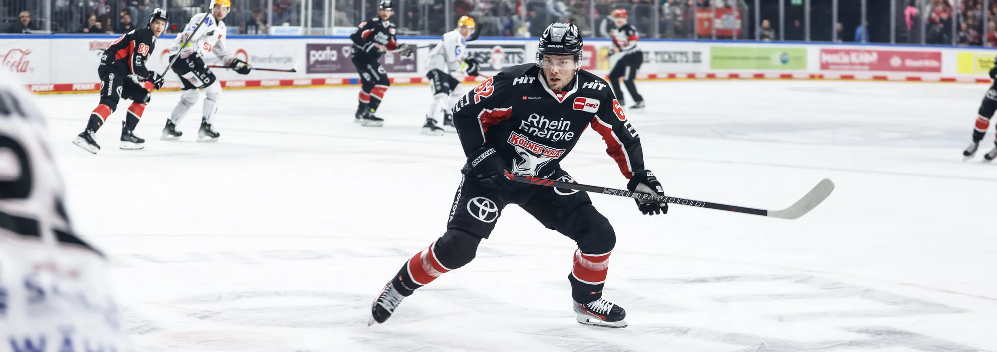 An ice hockey player in a black and red jersey skating on the ice with a hockey stick.