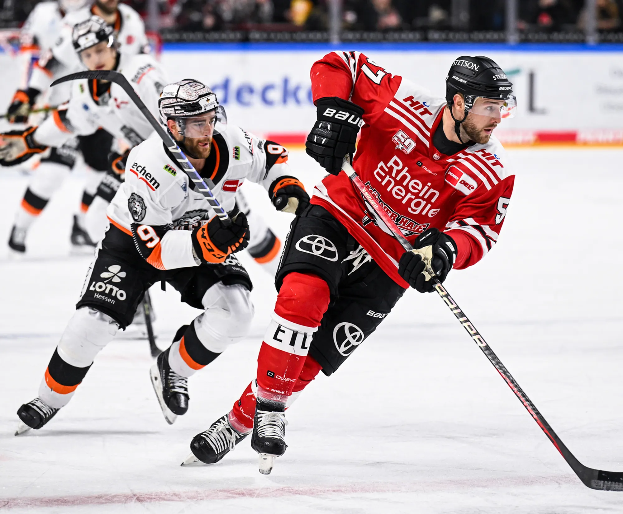 Two ice hockey players on the ice, one in a red jersey and the other in a white jersey with orange accents.