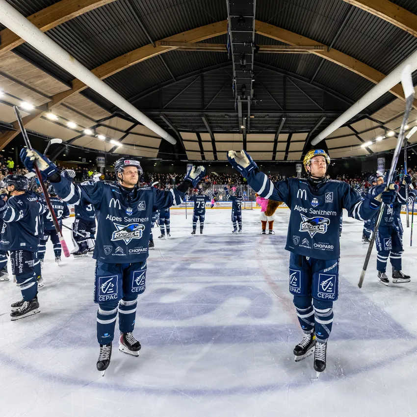 Two ice hockey players in blue jerseys standing on the ice, raising their sticks as a cheering crowd is visible in the background.