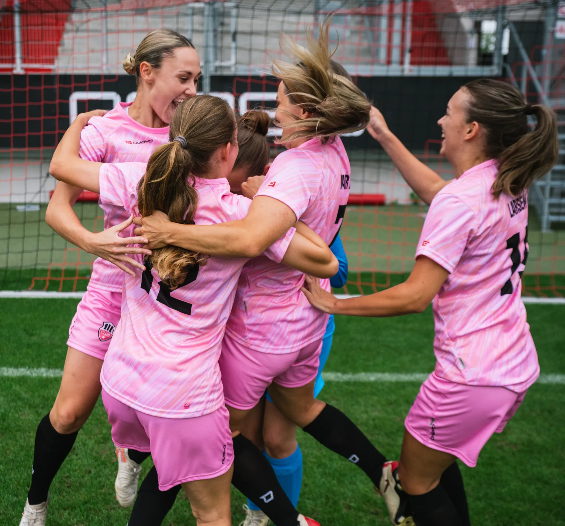 Five female players in pink jerseys joyfully celebrate together on the soccer field.