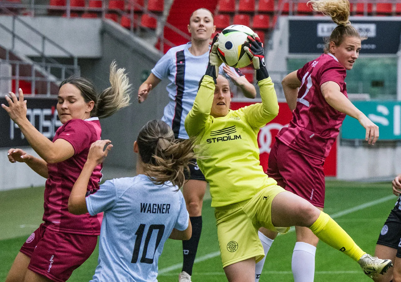 A goalkeeper in yellow sportswear catches the ball while players in various jerseys are engaged around her.