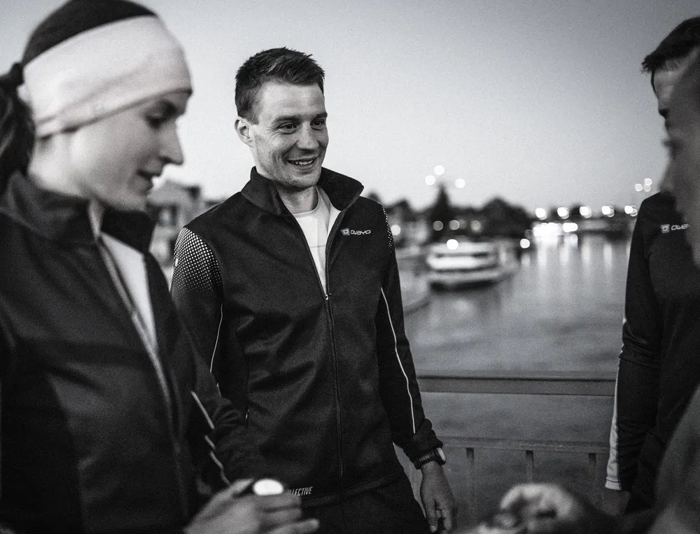 Two athletes in modern black jackets stand by a waterfront, engaging in conversation at dusk.