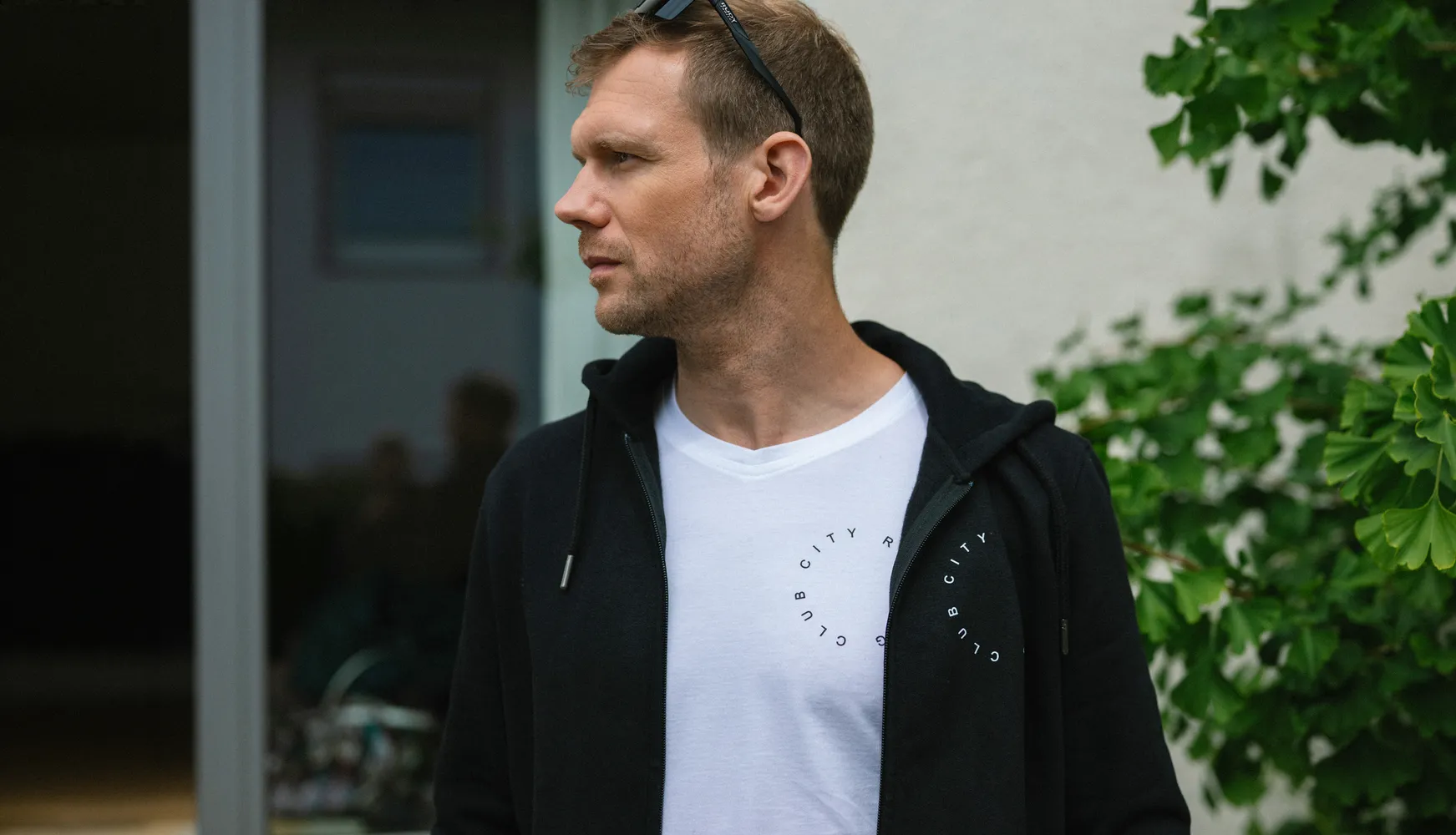 A man wearing a black hoodie over a white t-shirt with a circular print stands in an outdoor setting.
