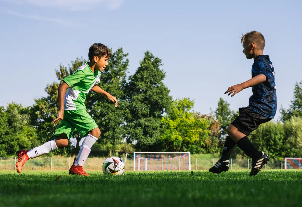 Two children are playing soccer on a green field, one wearing a green jersey with white accents and the other in a blue shirt, as they chase a ball.