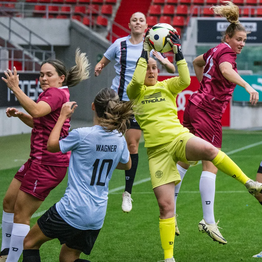 A goalkeeper in a yellow jersey catches the ball while players in various jerseys are active on the soccer field.