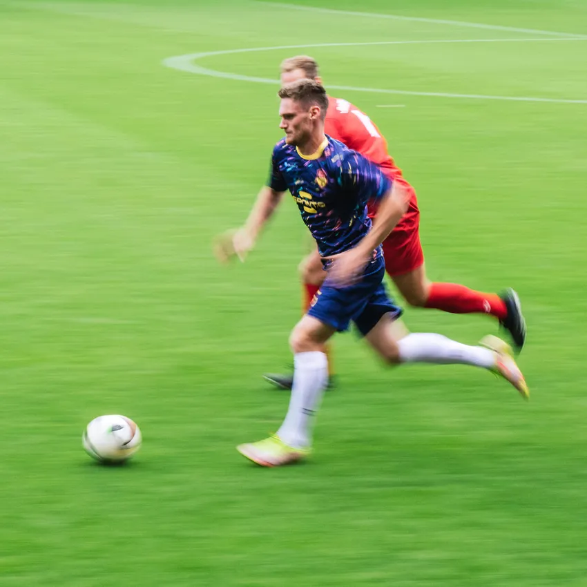 A football player in a colorful jersey sprints across the green field while another player pursues him.