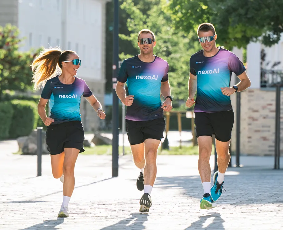 Three athletes, two men and one woman, are jogging in matching colorful running shirts and shorts on a paved path.