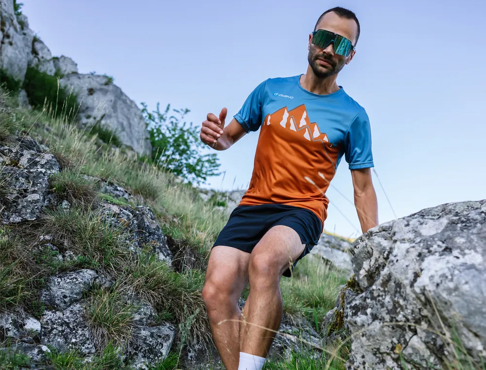 A man is running over a rocky path wearing a sporty blue and orange T-shirt and shorts.