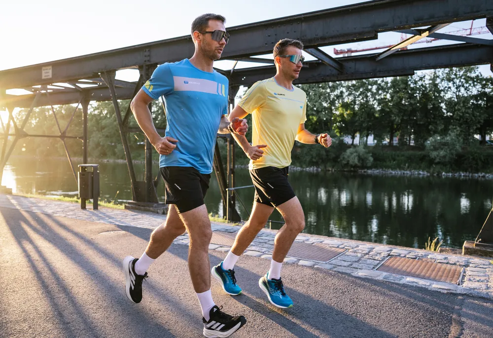 Two men in sportswear jog along a calm body of water as the sun rises.