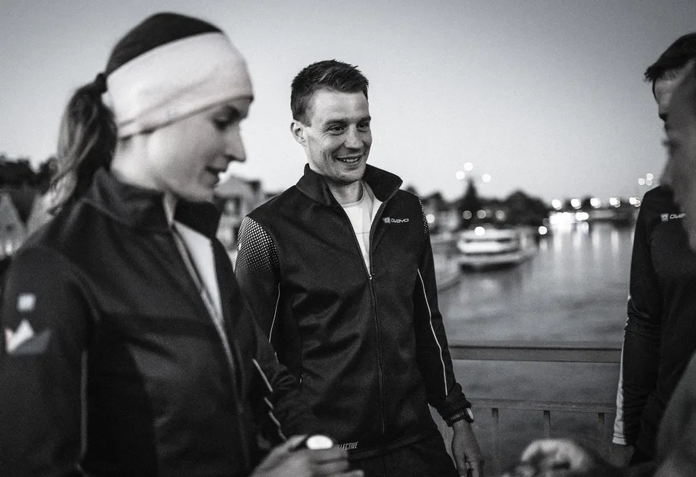 Two individuals are wearing sports jackets and chatting casually by a body of water during twilight.