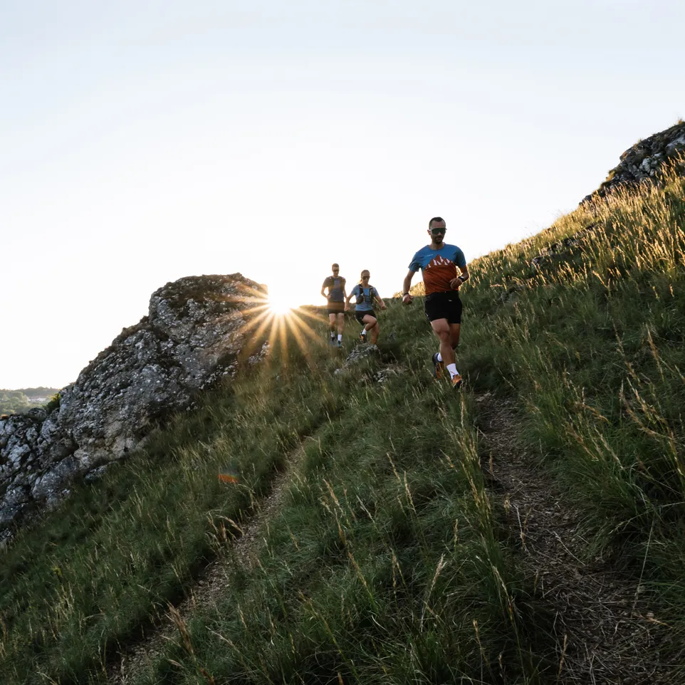 Three runners in athletic wear jog along a hilly path as the sun rises behind them.