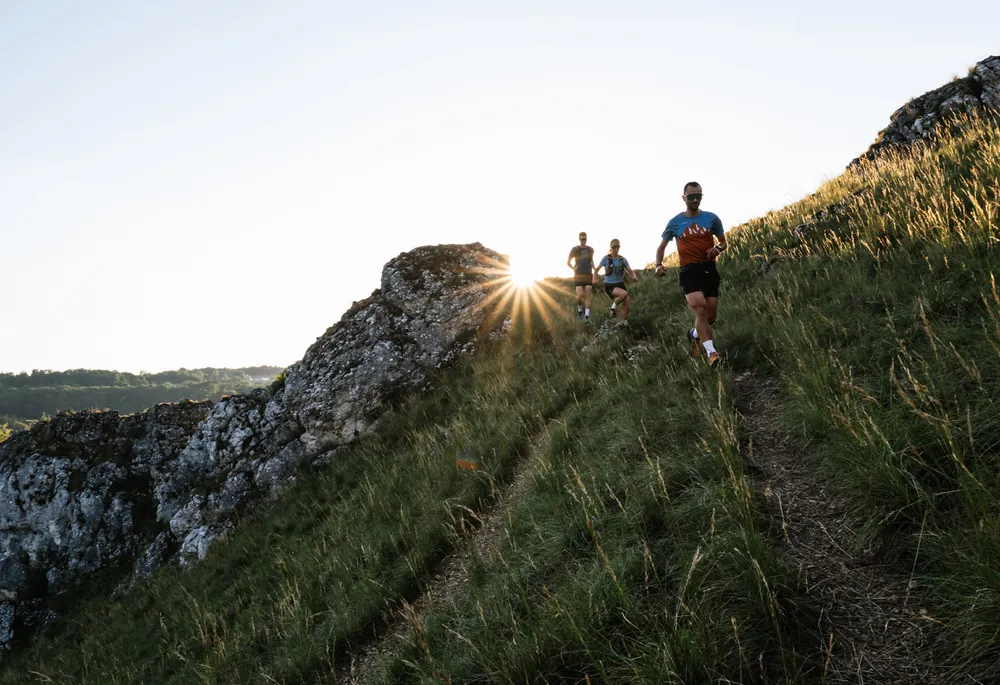 A group of three runners descending a hilly path, surrounded by green grass and rocks, with the sun rising in the background.