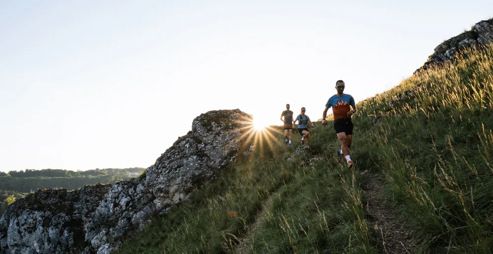Three runners in functional sportswear are running along a hilly path as the sun rises behind them.