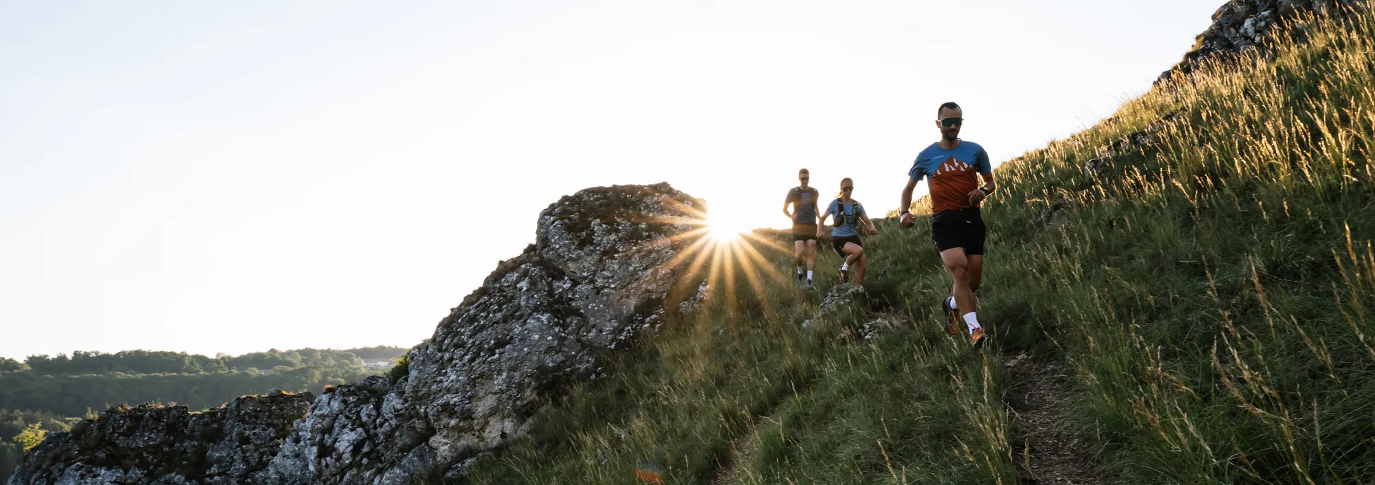 Three people in athletic clothing are running along a hilly path as the sun rises behind them.