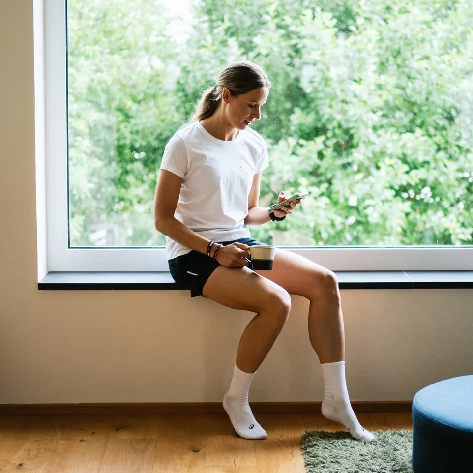 A woman sits on a windowsill, wearing a white t-shirt and black shorts, while holding a smartphone and enjoying a cup of coffee.