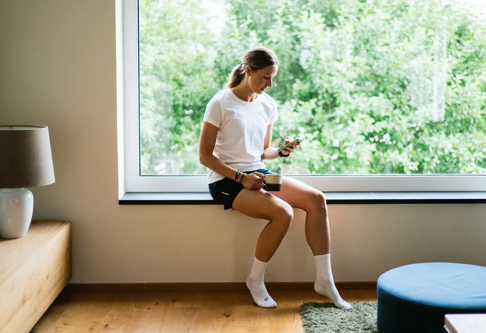 A woman sits on a windowsill holding a phone in one hand and a cup in the other, dressed in a white shirt and black shorts.