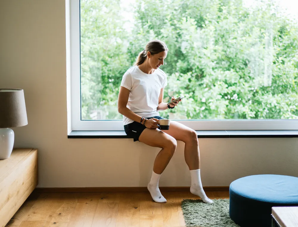 A woman sits on a windowsill, looking at her phone while holding a cup of coffee, dressed in a white t-shirt and black shorts.
