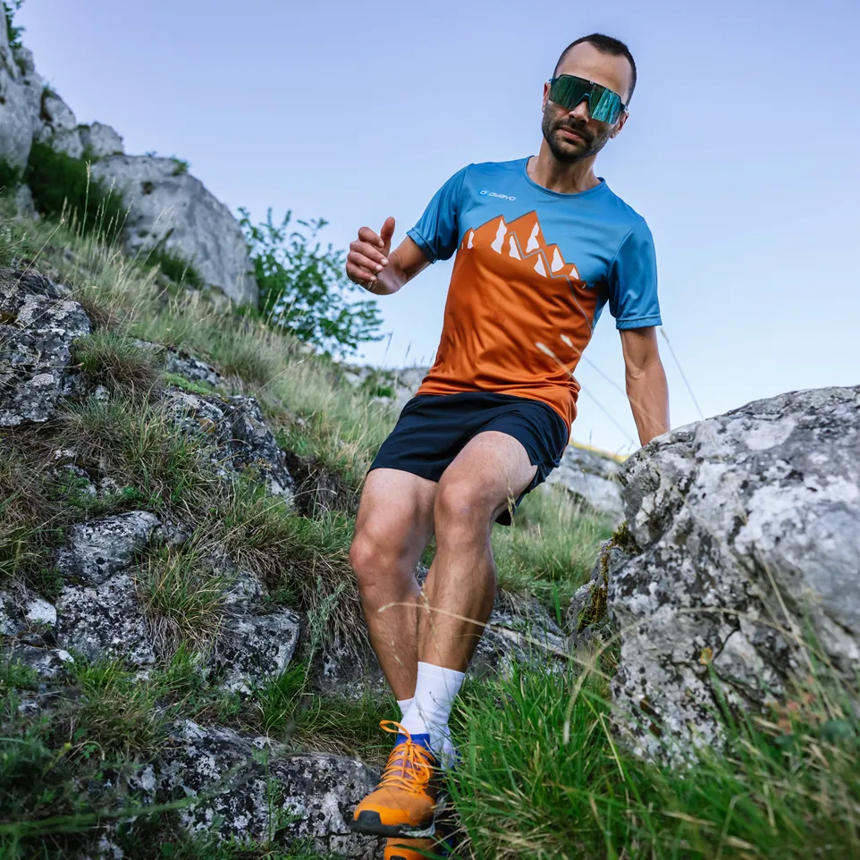A man is running on a rocky path, wearing a blue and orange short-sleeved sports shirt with mountain design and black shorts.