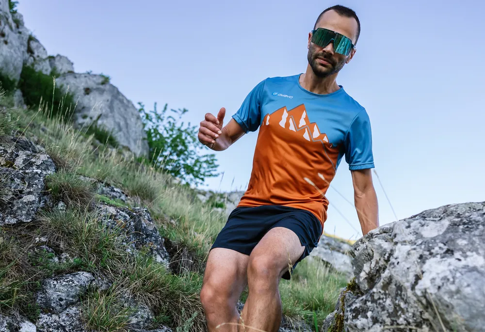A man is running on a rocky path wearing a colorful sports shirt with a mountain design.
