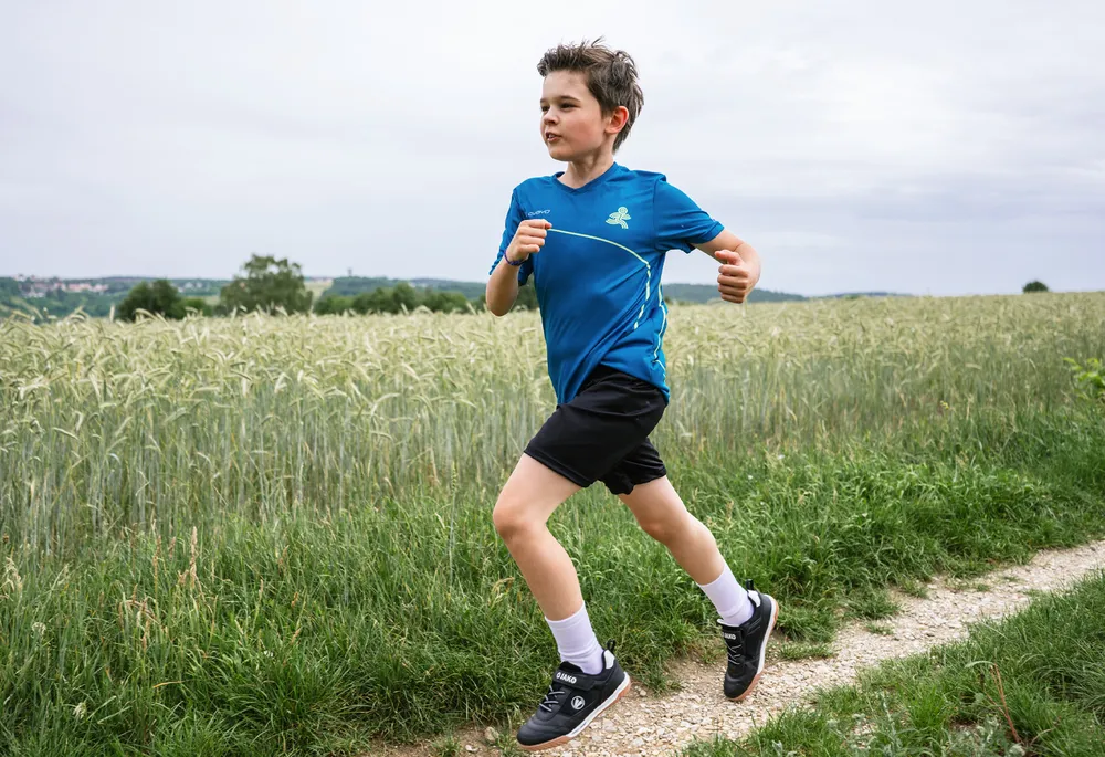 A boy in a blue sports shirt and black shorts runs along a path through a field.