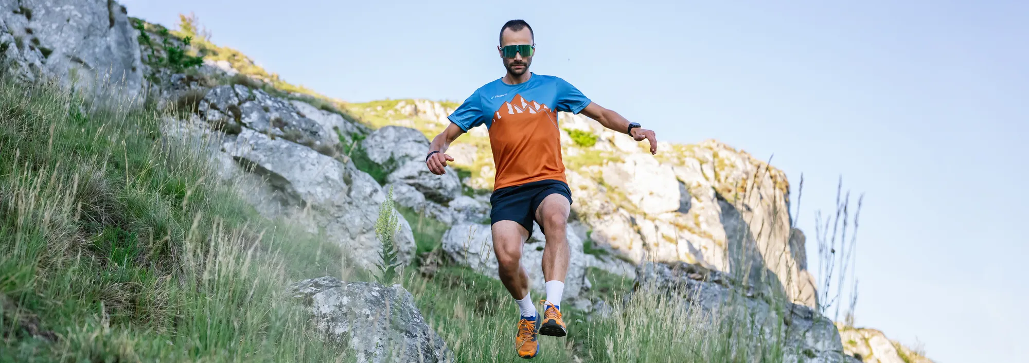 A male runner in an orange and blue shirt and shorts, running down a rocky slope.