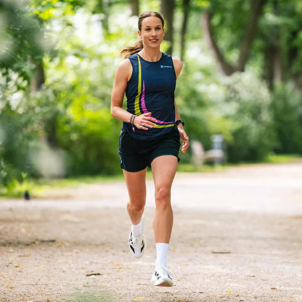 A sporty woman runs on a path in a park, wearing a sleeveless dark blue top with colorful accents and shorts.