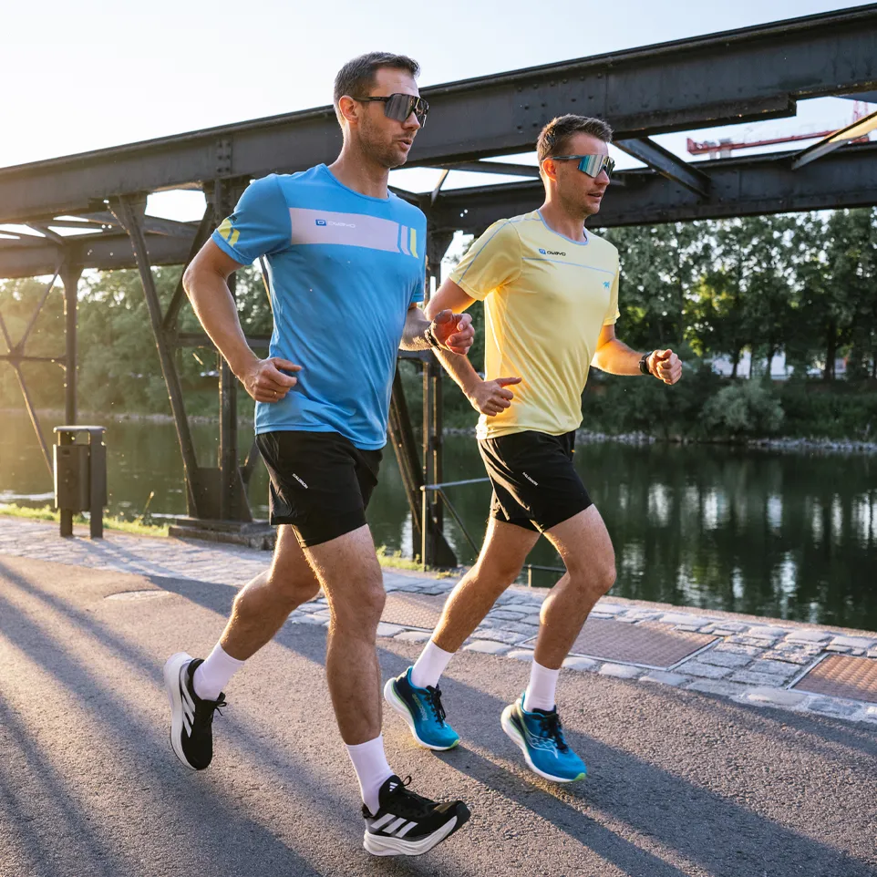 Two men are jogging on a path beside a body of water, one wearing a blue t-shirt and the other a yellow t-shirt.