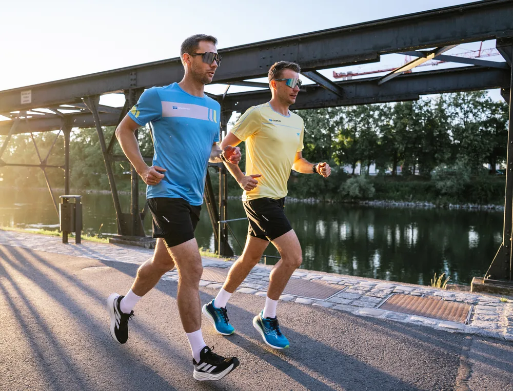 Two men are jogging by the water's edge, one wearing a blue t-shirt and the other a yellow one, both in shorts.