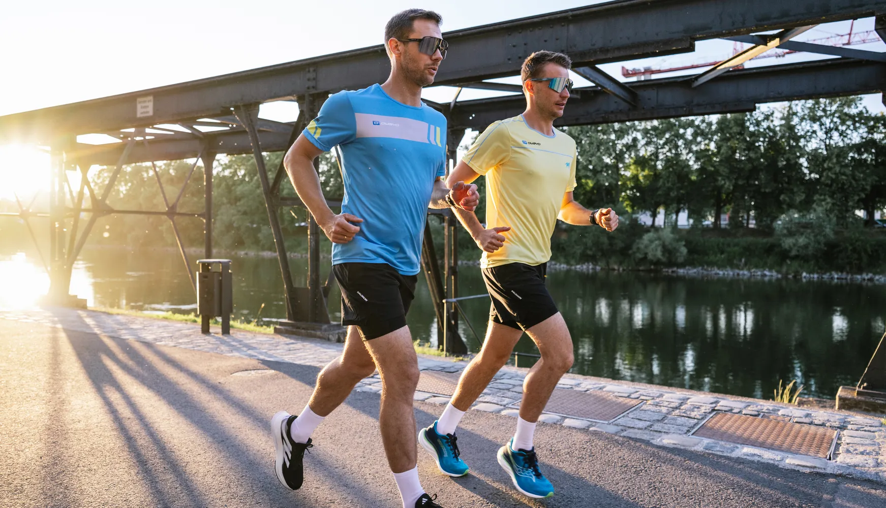 Two men in athletic running gear, one wearing a blue t-shirt and the other a yellow t-shirt, jog along the bank of a waterway at sunset.