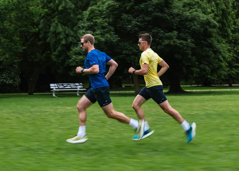 Two men in athletic wear are running on a green field, one wearing a blue shirt and the other a yellow shirt.