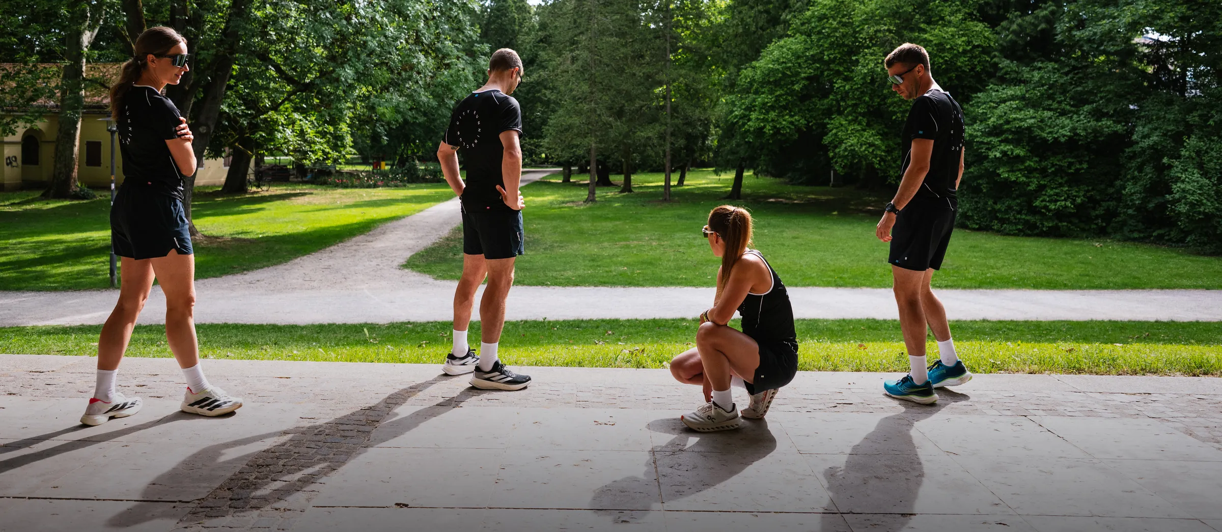 Four individuals in athletic clothing are standing and sitting in a green park landscape.