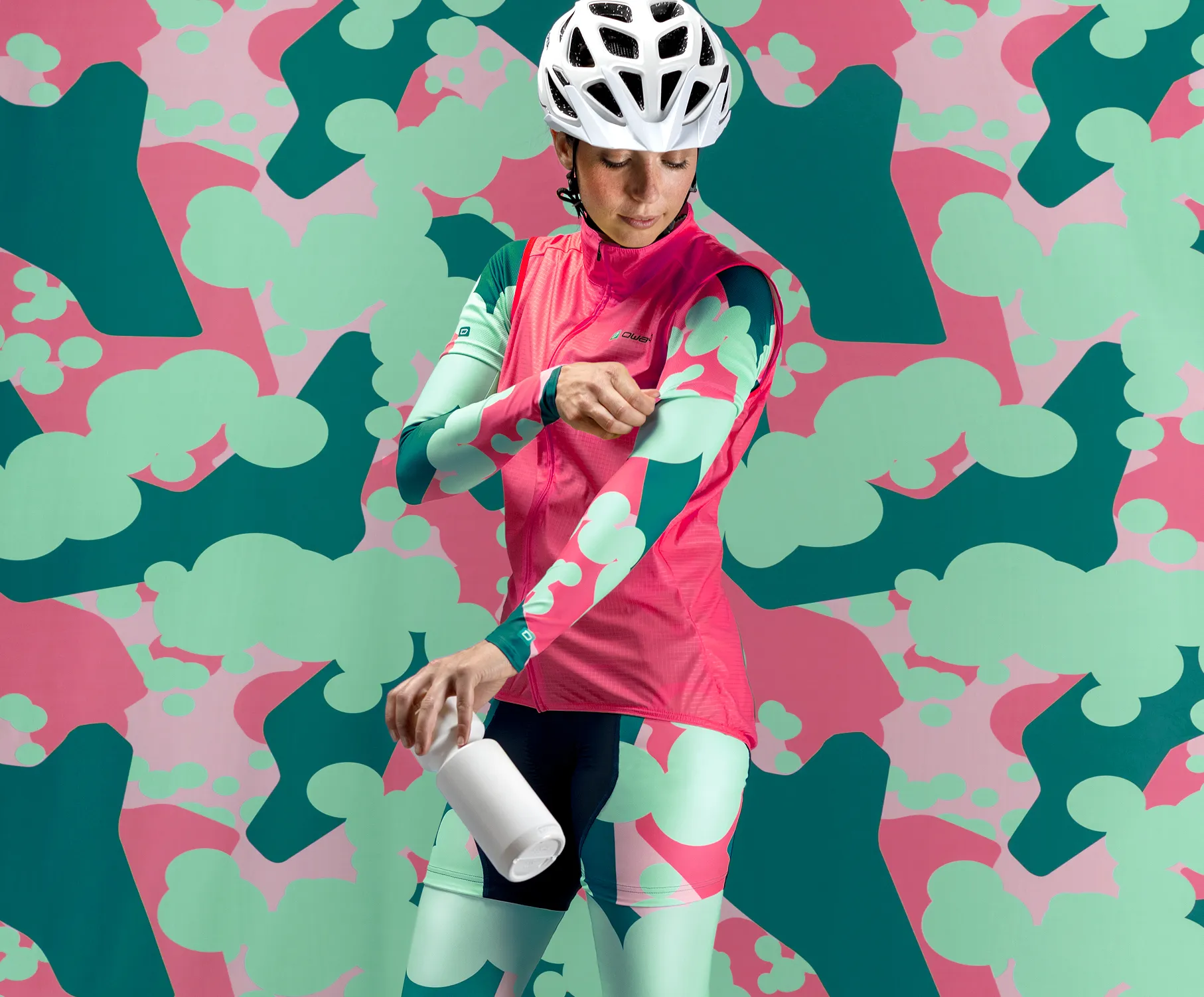 A cyclist in a custom pink and green sportswear with an abstract pattern, adjusting her sleeves while holding a white bottle, against a colorful background with the same pattern.