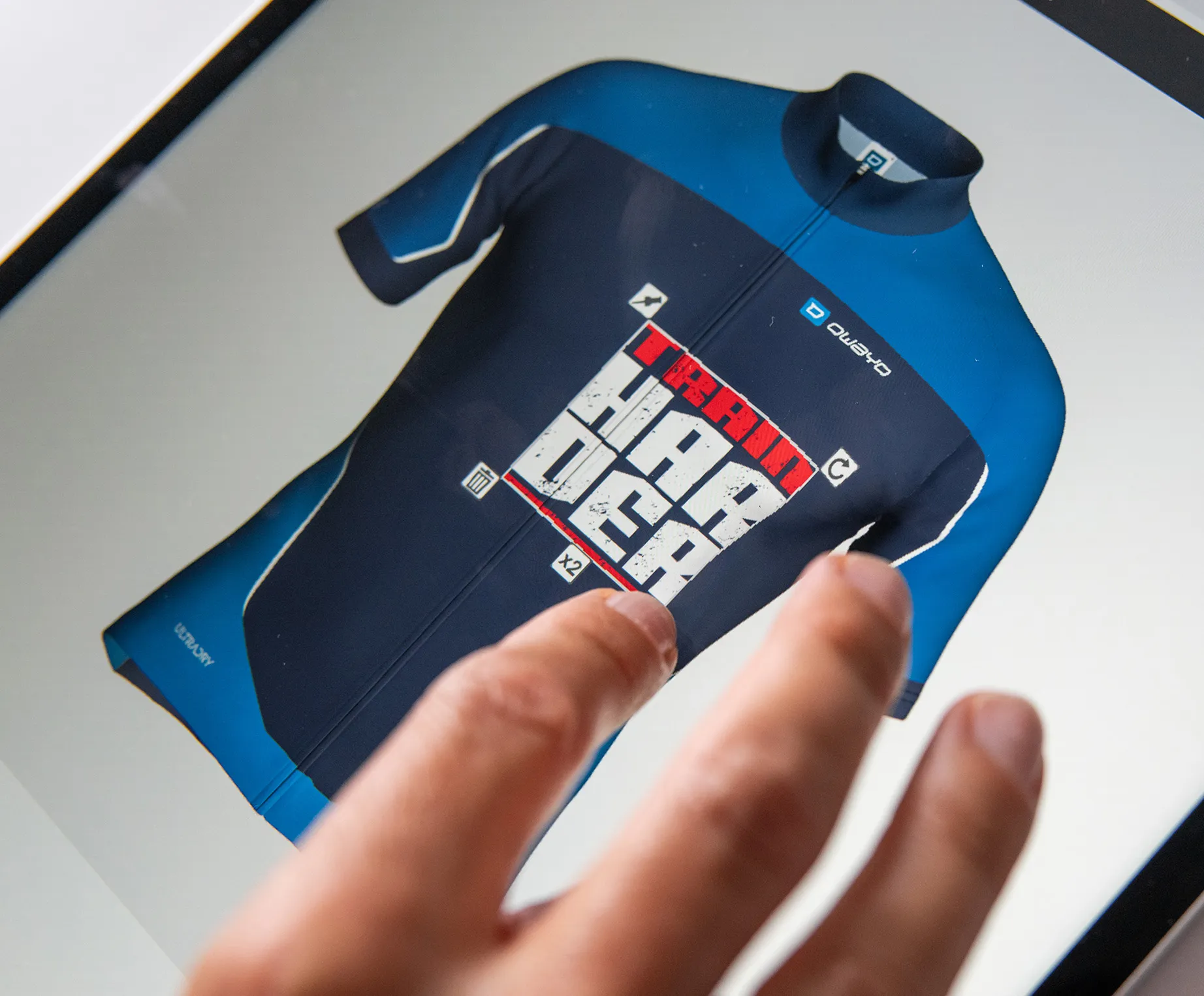 A hand touches a screen displaying a customized dark blue and blue sports jersey with the text 'TRAIN HARDER' and the owayo logo.