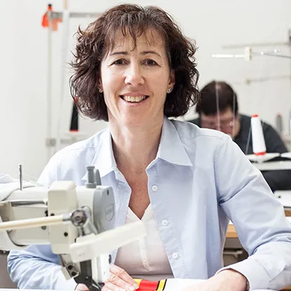 A woman smiles at the camera while working at a sewing machine in a workshop, with another person visible in the background also working at a sewing machine.