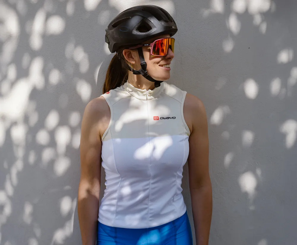 A woman wearing a white sleeveless cycling jersey with the owayo logo, a bicycle helmet, and sunglasses, leaning against a wall with leaf shadows.