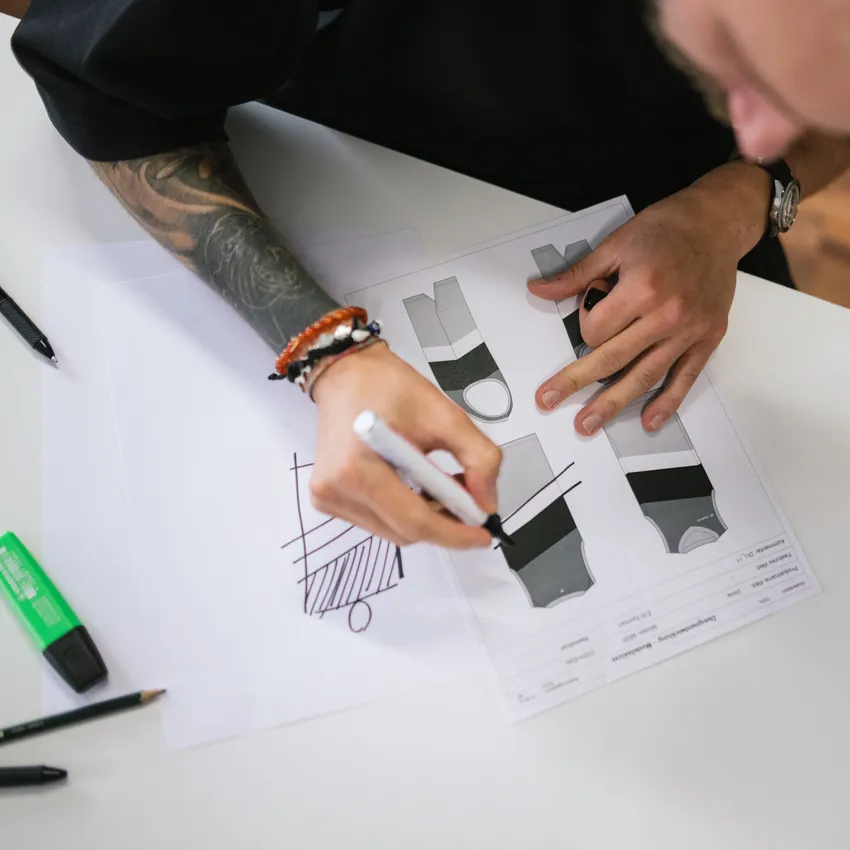 A person with a tattooed arm is sketching designs for sportswear on paper, surrounded by writing utensils.