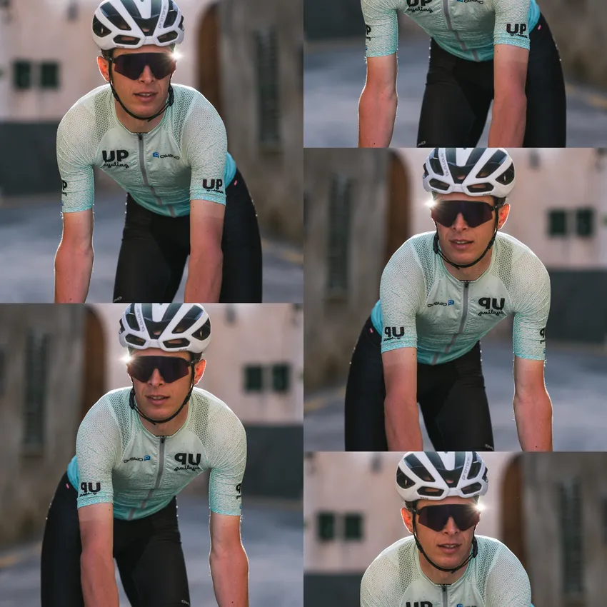 A cyclist wearing a light blue short-sleeve jersey and black shorts rides on a sunny street.