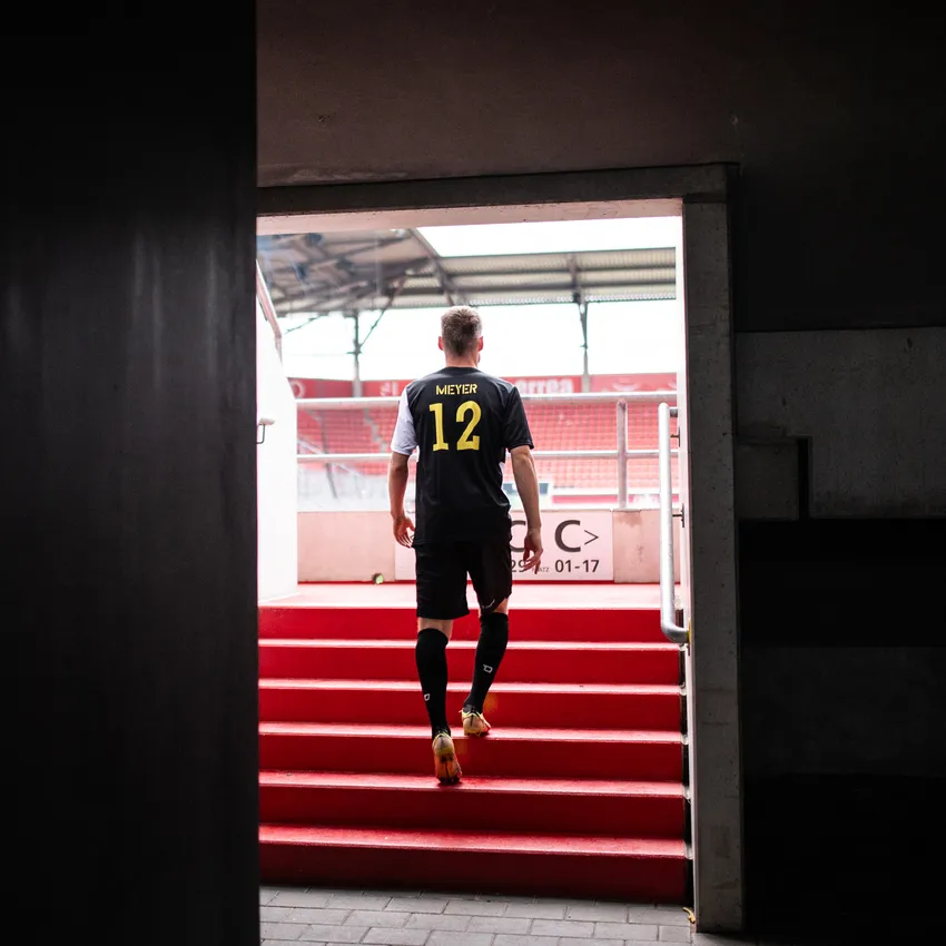 A football player in a black jersey with the number 12 ascends the red steps of a stadium.
