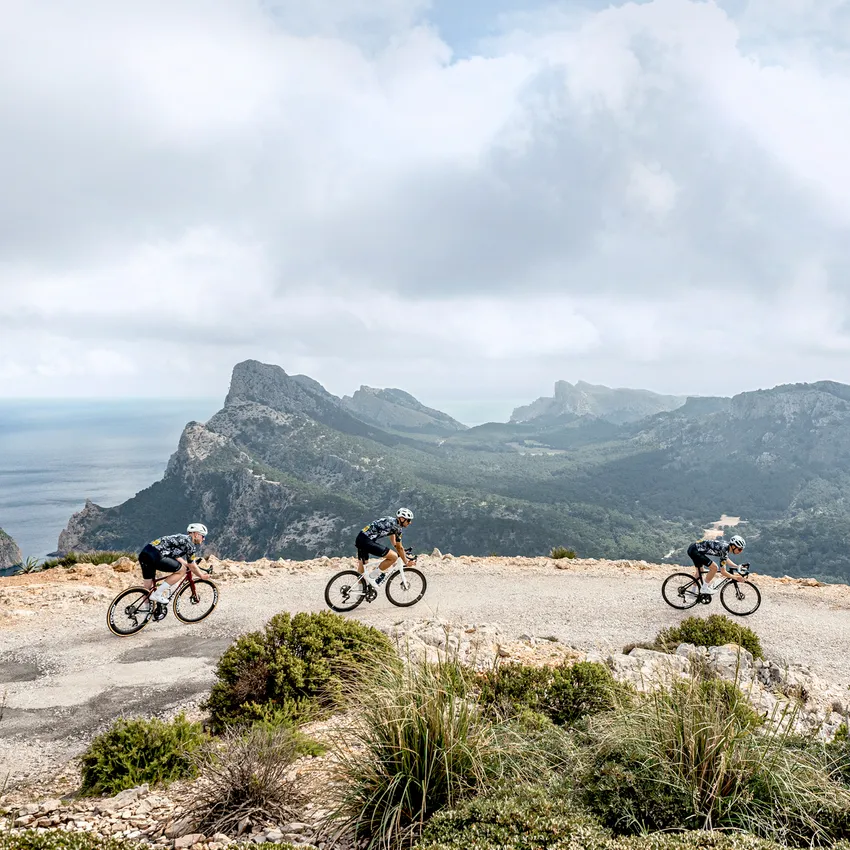 Three cyclists in sportswear ride on a winding road with a mountainous landscape and sea in the background.