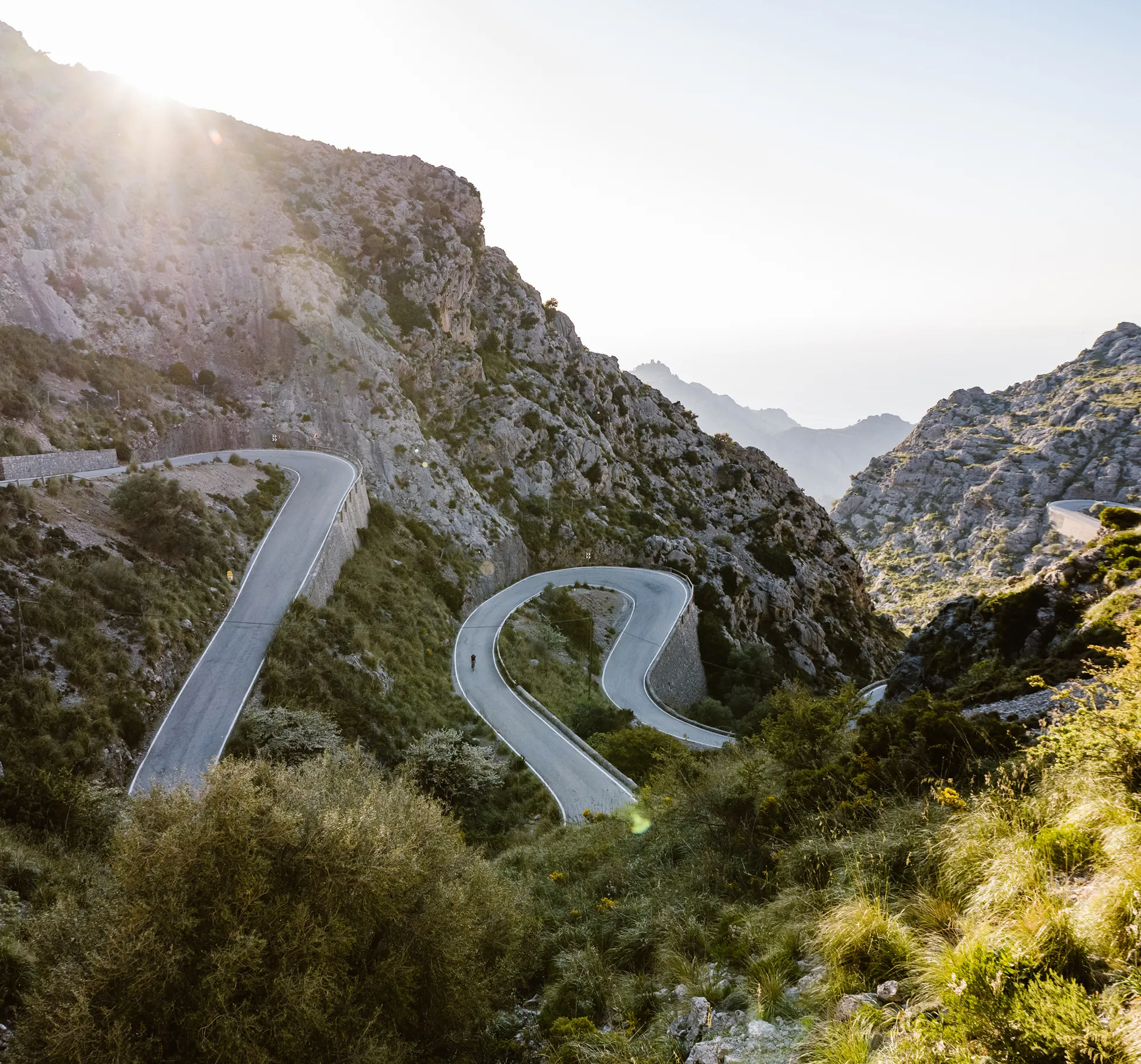 A person rides a bicycle along a winding mountain road that snakes through a rocky landscape, with sunlight shining from above.