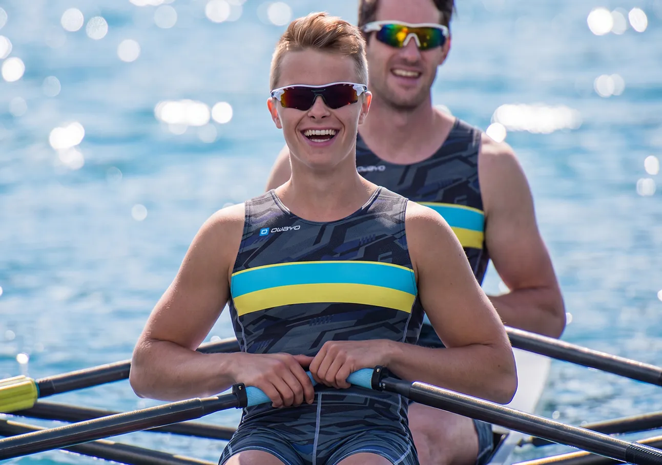 Two cheerful rowers in athletic suits with blue and yellow stripes sitting in a boat on sparkling water.