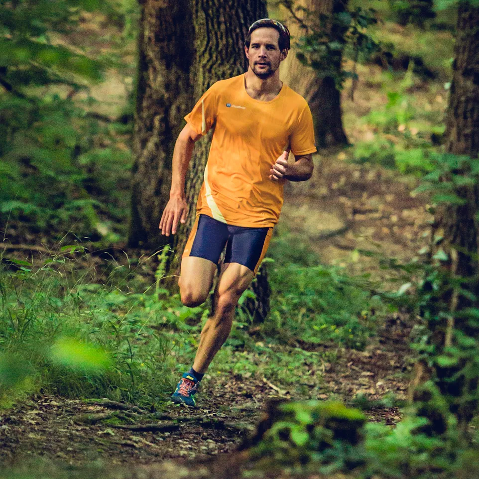 A man running on a forest trail wearing an orange running jersey and dark blue running shorts.