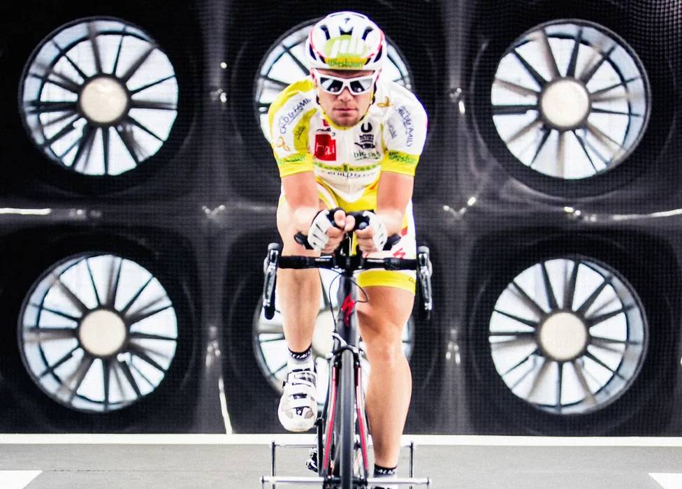 A cyclist in a yellow and white jersey and helmet rides a stationary bike in front of a background with large fans.