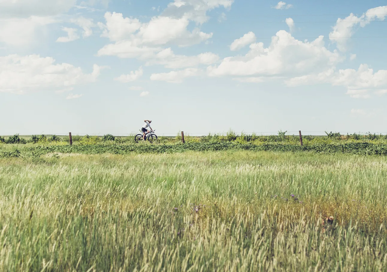 A person rides a bicycle through a green field under a blue sky with white clouds.