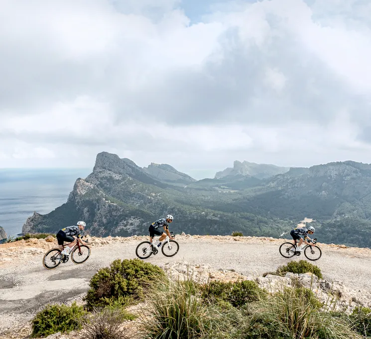 Three cyclists in sportswear ride on a road along a coastal landscape with mountains and sea in the background.